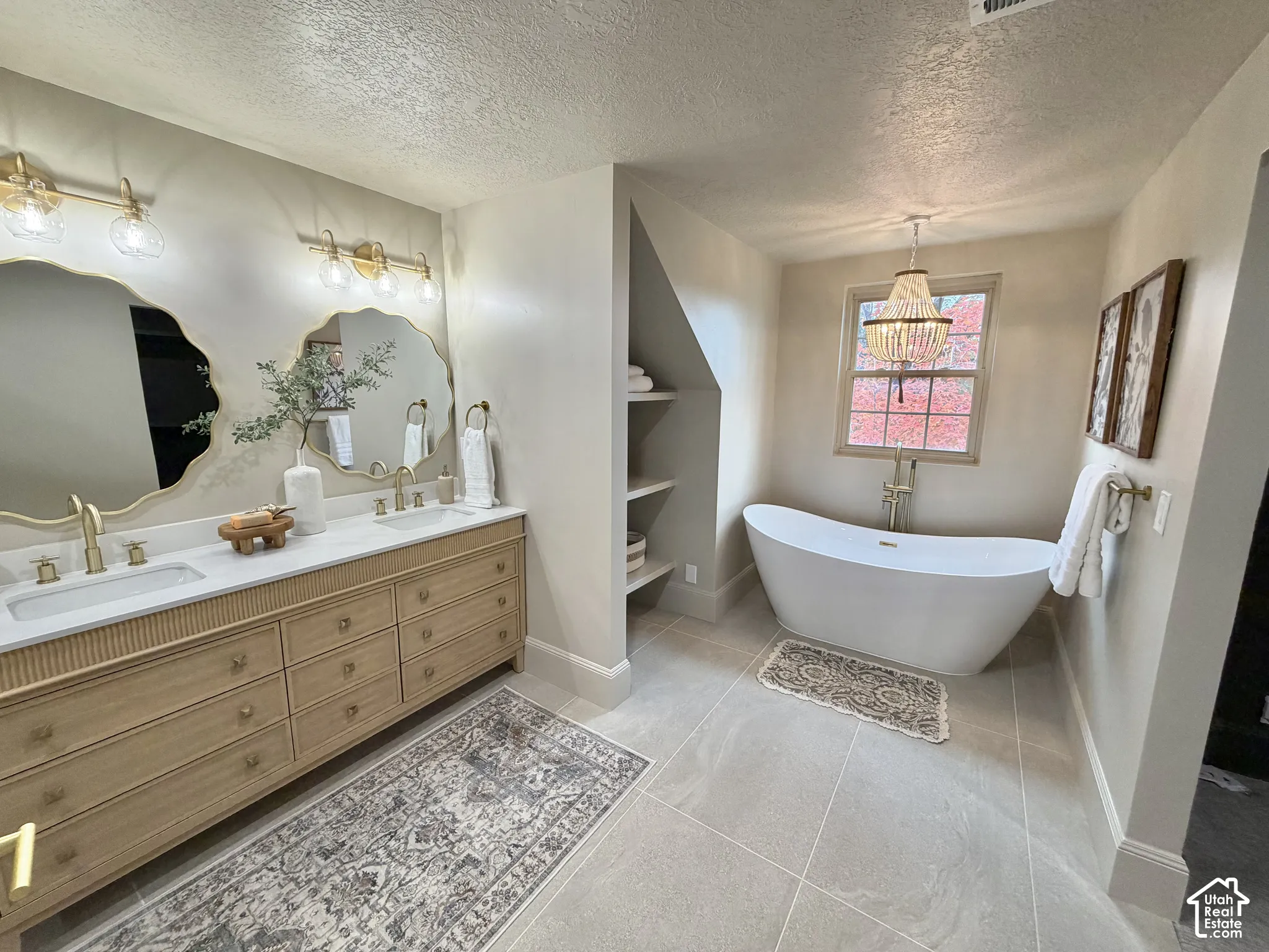 Dual-vanity cabinet with light wood finish and light-colored countertop, wall-mounted scalloped mirrors, freestanding soaking tub with floor-mounted faucet, and built-in shelving