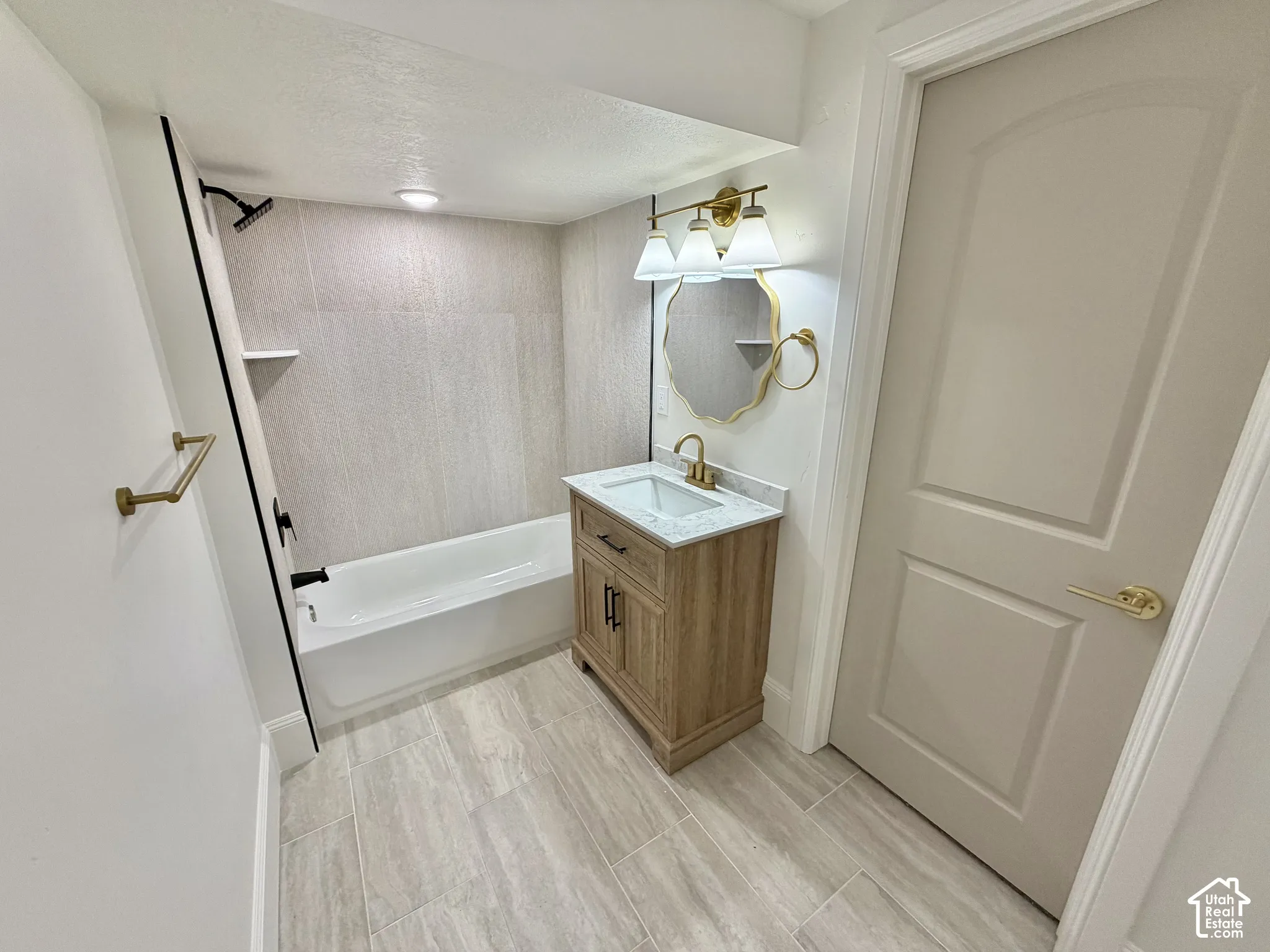 Bathroom featuring a vanity with a light-finish wood cabinet and stone-look countertop, a bathtub with full-height tile surround, brass-finish hardware, and tile flooring
