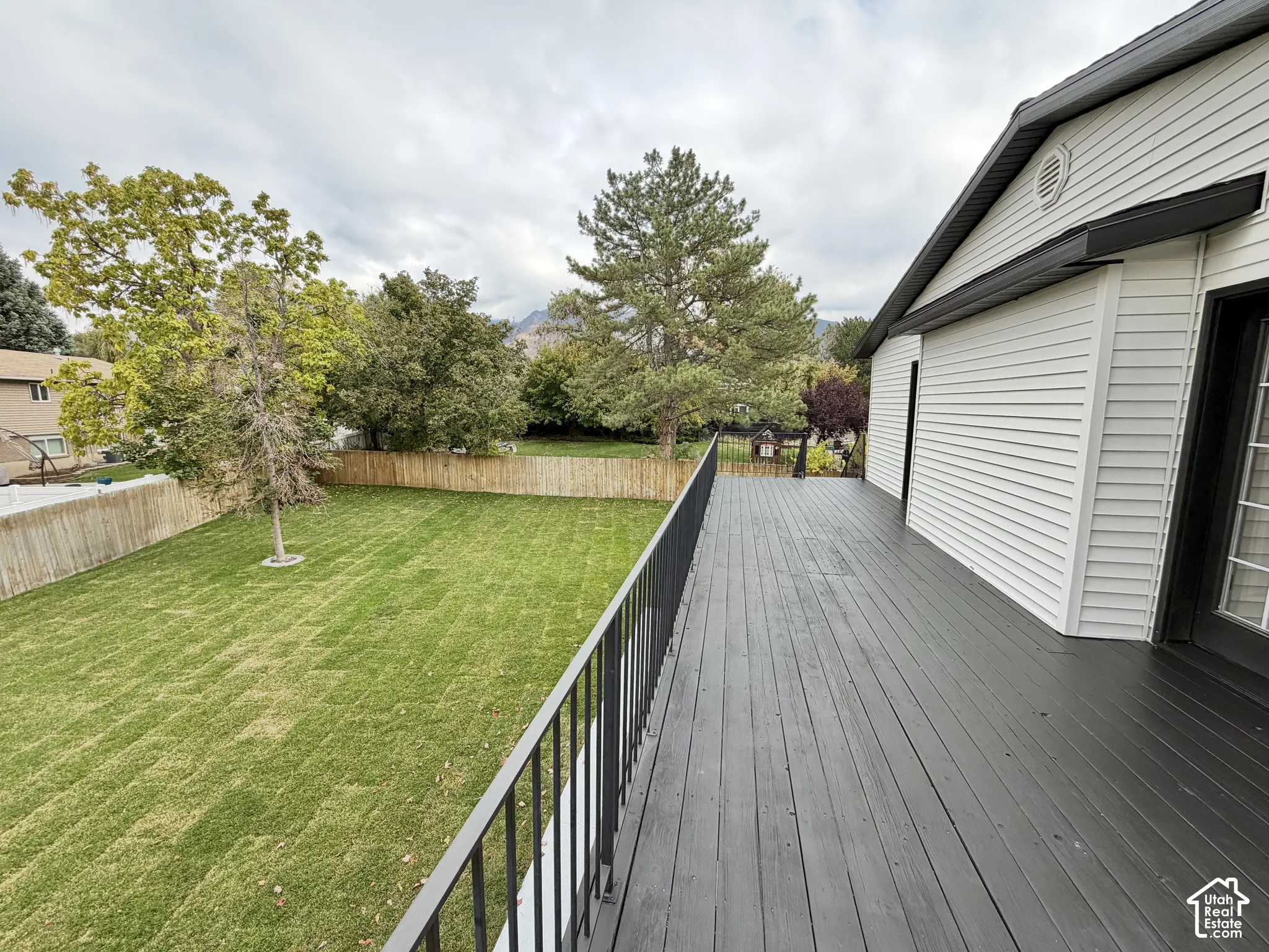 Elevated deck with dark-stained wood flooring and metal railings, overlooking a fenced backyard featuring a level lawn and mature trees