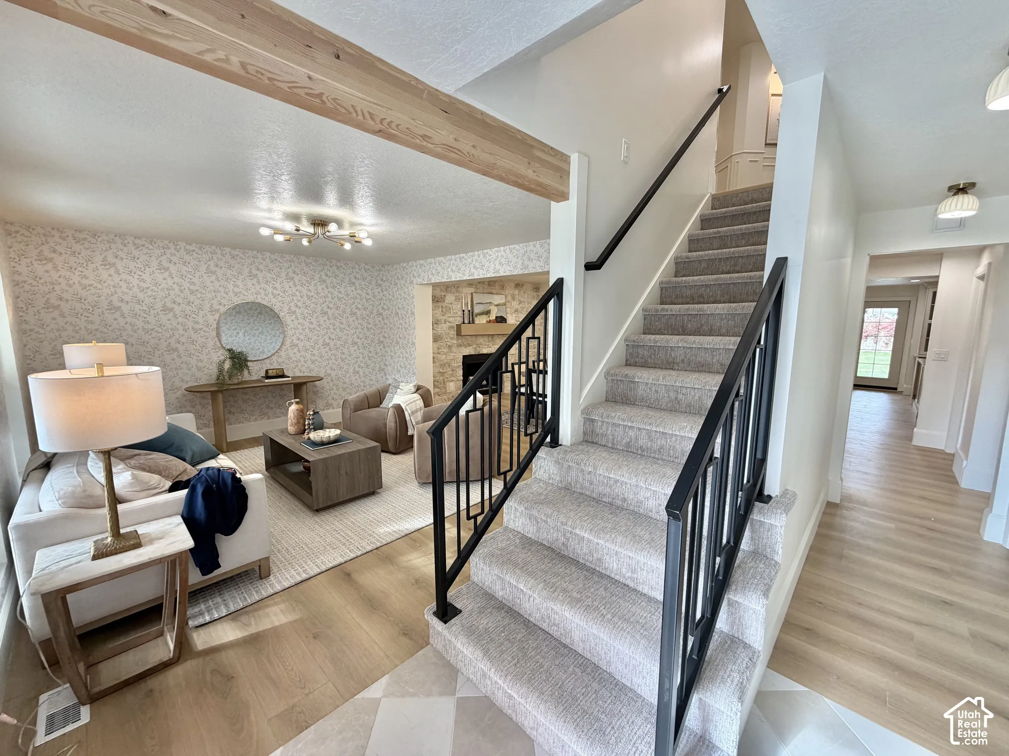 Interior entryway featuring a staircase with a geometric matte black railing, exposed natural wood ceiling beam, stone-faced fireplace, and wood-finish flooring