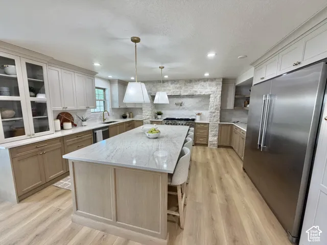 Kitchen featuring light wood-finish cabinetry, a large central island with stone-surface countertop, floor-to-ceiling stone range surround, stainless steel refrigerator, and pendant lighting