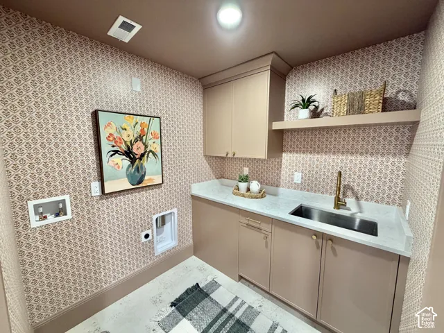 Utility room featuring custom cabinetry, light-toned stone-finish countertops, an undermount stainless steel sink with a brass-finish faucet, and a wall-mounted floating shelf