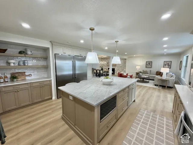 Kitchen featuring a central island with stone countertop, stainless steel double-door refrigerator, custom wood-finish cabinetry, recessed lighting, and floating shelves with tile backsplash