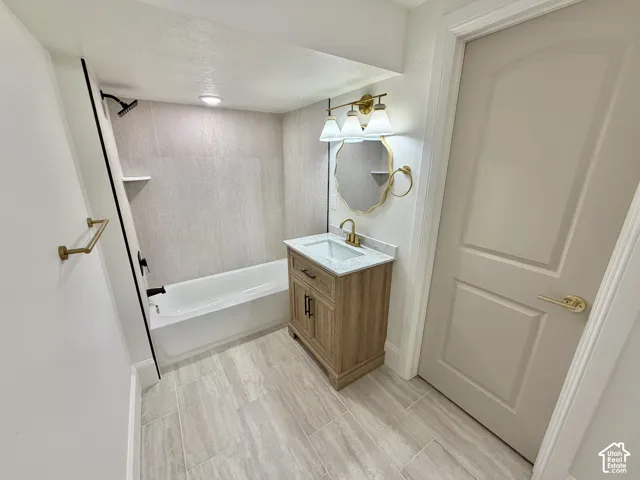 Bathroom featuring a vanity with a light-finish wood cabinet and stone-look countertop, a bathtub with full-height tile surround, brass-finish hardware, and tile flooring