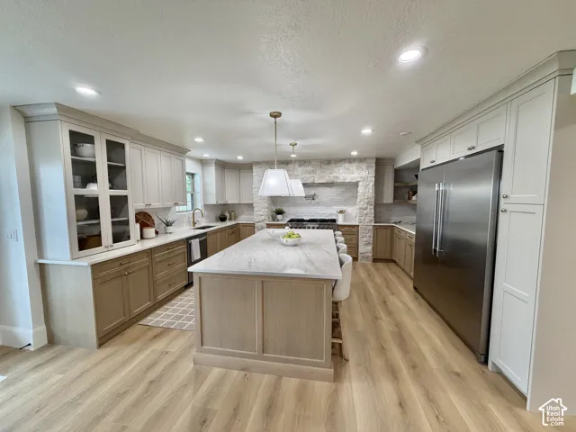 Kitchen featuring a large central island with light-toned stone-finish countertop, custom two-tone cabinetry, recessed lighting, stainless steel refrigerator, and a decorative stone-clad range hood