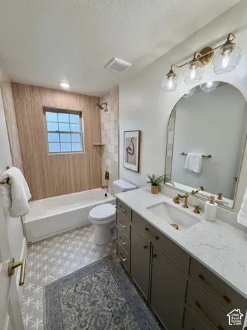 Bathroom featuring a vanity with quartz countertop, arched wall mirror, three-light brass fixture, patterned tile flooring, and tub surround with wood-finish tile accents