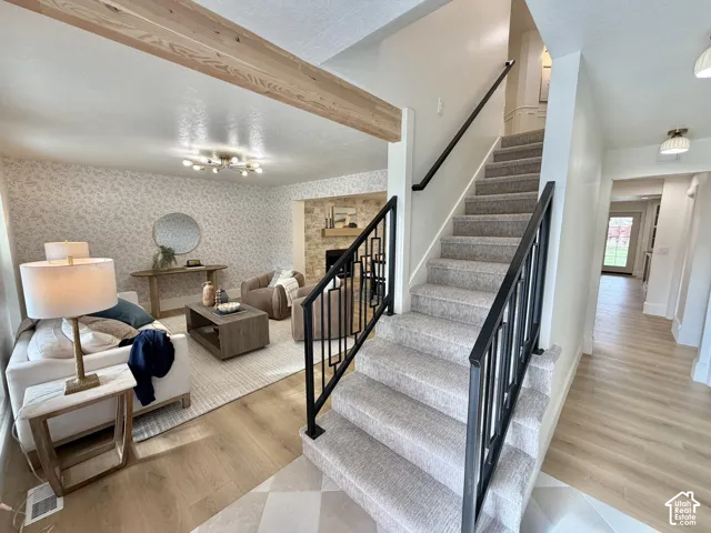 Interior entryway featuring a staircase with a geometric matte black railing, exposed natural wood ceiling beam, stone-faced fireplace, and wood-finish flooring