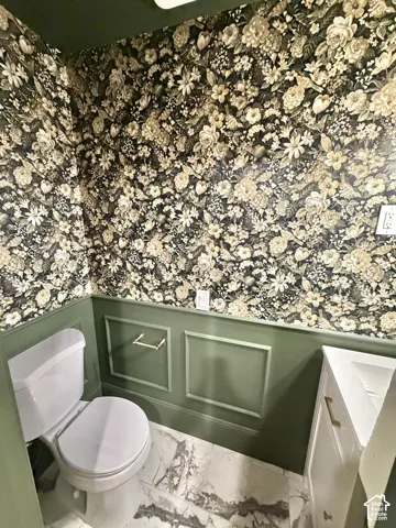 Green paneled wainscoting, floral patterned wallpaper, white vanity with gold hardware, and stone-finish flooring