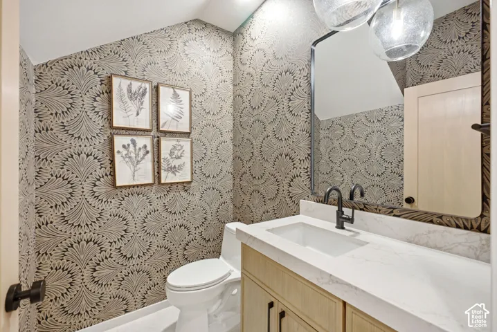 Bathroom featuring a wood-finish vanity with a stone countertop, an integrated rectangular sink, matte black plumbing fixtures, and patterned wallpaper