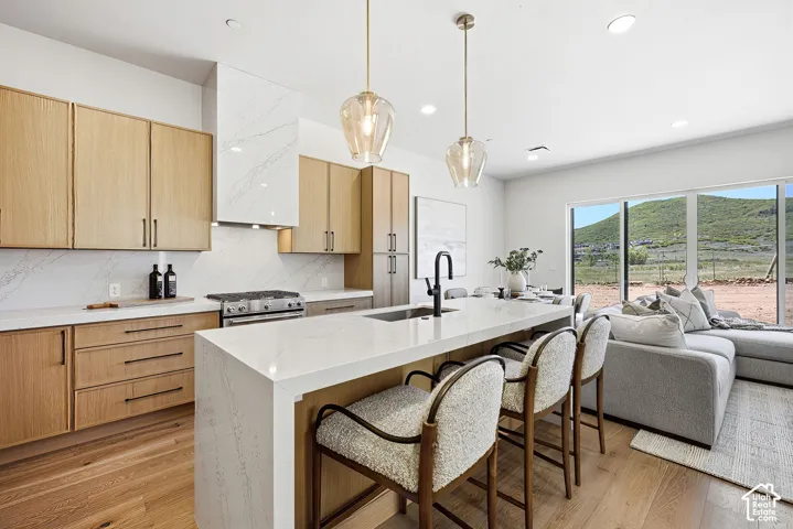 Kitchen featuring light wood-finish cabinetry, quartz countertops, a matching full-height backsplash, a waterfall edge island, and pendant lighting