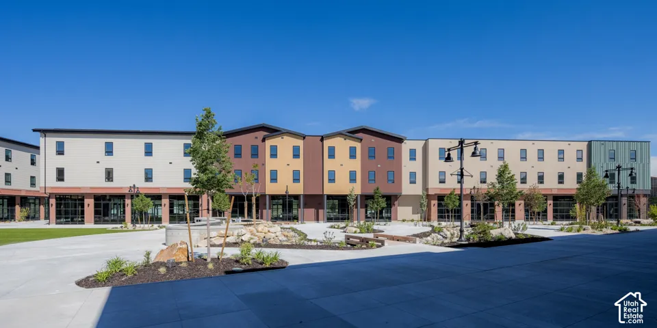 Multi-story exterior featuring multi-toned horizontal siding, brick masonry detailing, ground-level glass storefronts, landscaped rock gardens, and concrete plaza paving