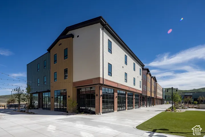 Multi-story exterior featuring a mix of horizontal siding, vertical metal paneling, and brick masonry, complemented by ground-level floor-to-ceiling glass storefronts, manicured synthetic lawn, and overhead string lighting