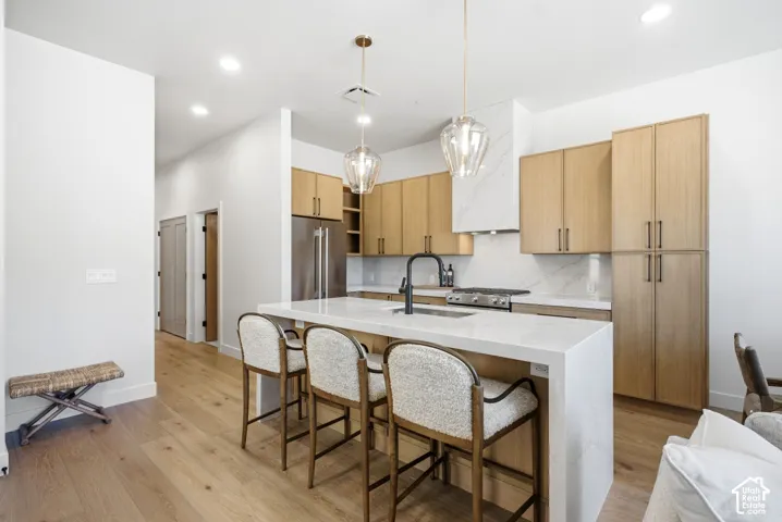 Kitchen featuring light wood-finish cabinetry, a waterfall-edge quartz island with an undermount sink, a matte black faucet, and glass pendant lighting