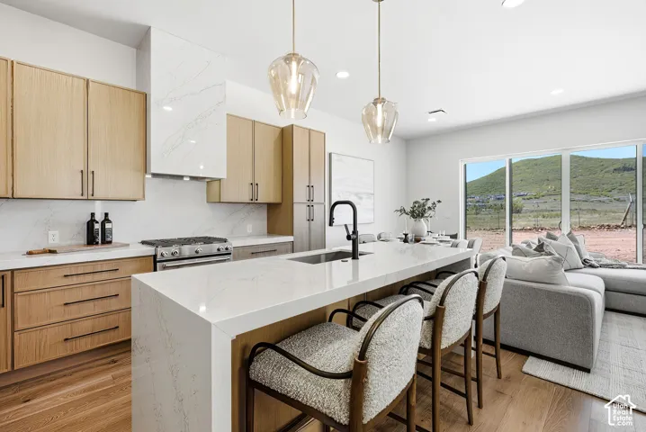 Kitchen featuring wood-finish cabinetry, stone-surface countertops with waterfall edge, full-height stone-surface backsplash, custom range hood, and wood-finish flooring