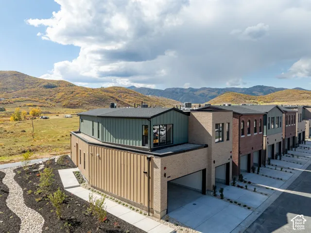 Contemporary multi-story exterior featuring tan vertical metal siding, green metal upper-level siding, brick masonry, and attached multi-car garages, set against a mountain backdrop