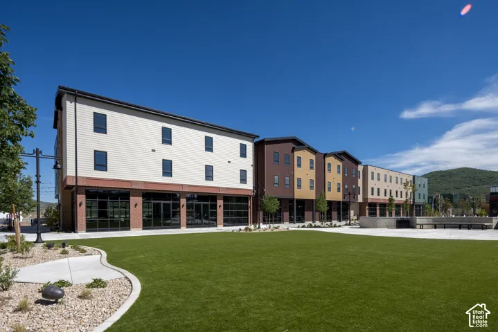 Multi-story mixed-use exterior featuring composite siding, brick masonry, ground-level glass storefronts, and an expansive artificial turf plaza