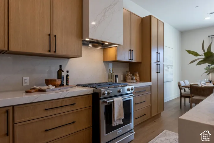Flat-panel wood-finish cabinetry, engineered stone countertops with matching full-height backsplash, stainless steel gas range, and integrated custom-cased range hood