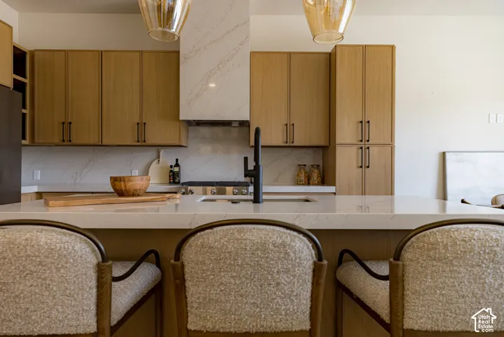 Kitchen featuring light-finish wood cabinetry, quartz countertops with full-height backsplash, a matching quartz range hood, matte black faucet, and pendant lighting