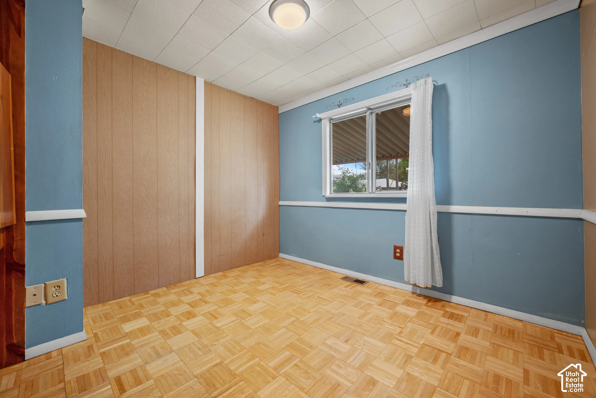 Room featuring wood-finish parquet flooring, paneled wall accents, a grid-patterned drop ceiling, and a central flush-mount light fixture