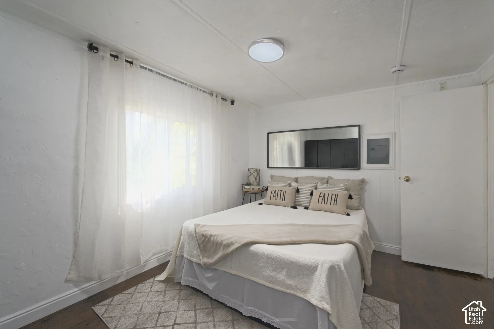 Room featuring wood-finish flooring, white walls, a flush-mount ceiling light fixture, a horizontal wall-mounted mirror, and a window with a curtain rod