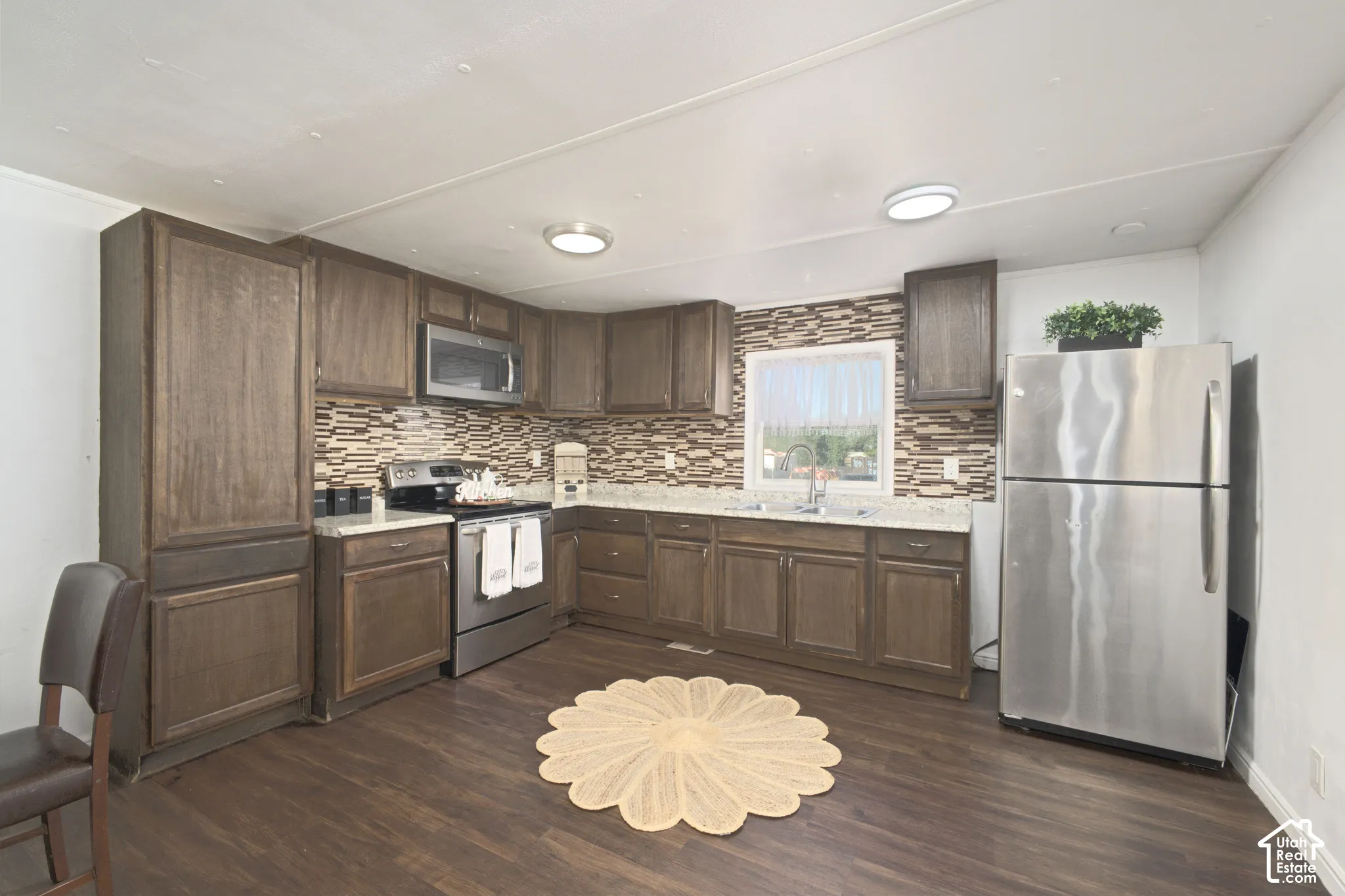 Kitchen featuring dark wood-finish cabinetry, stone-texture tile backsplash, wood-finish flooring, stainless steel appliances, and solid-surface countertops