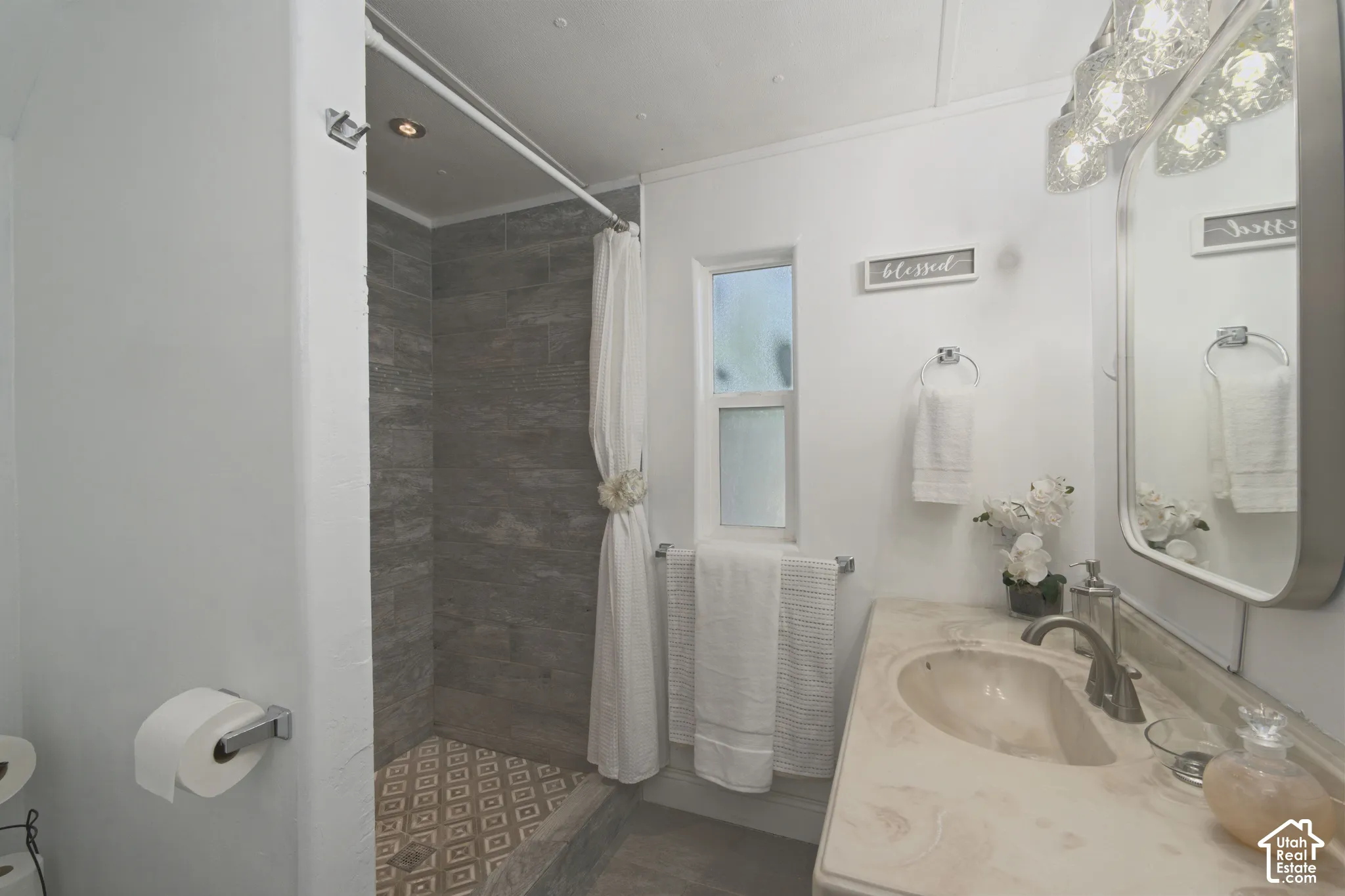 Bathroom featuring a shower stall with grey tile surround and decorative geometric tile flooring, solid-surface vanity countertop with an integrated sink, and an arched wall-mounted mirror with integrated overhead lighting