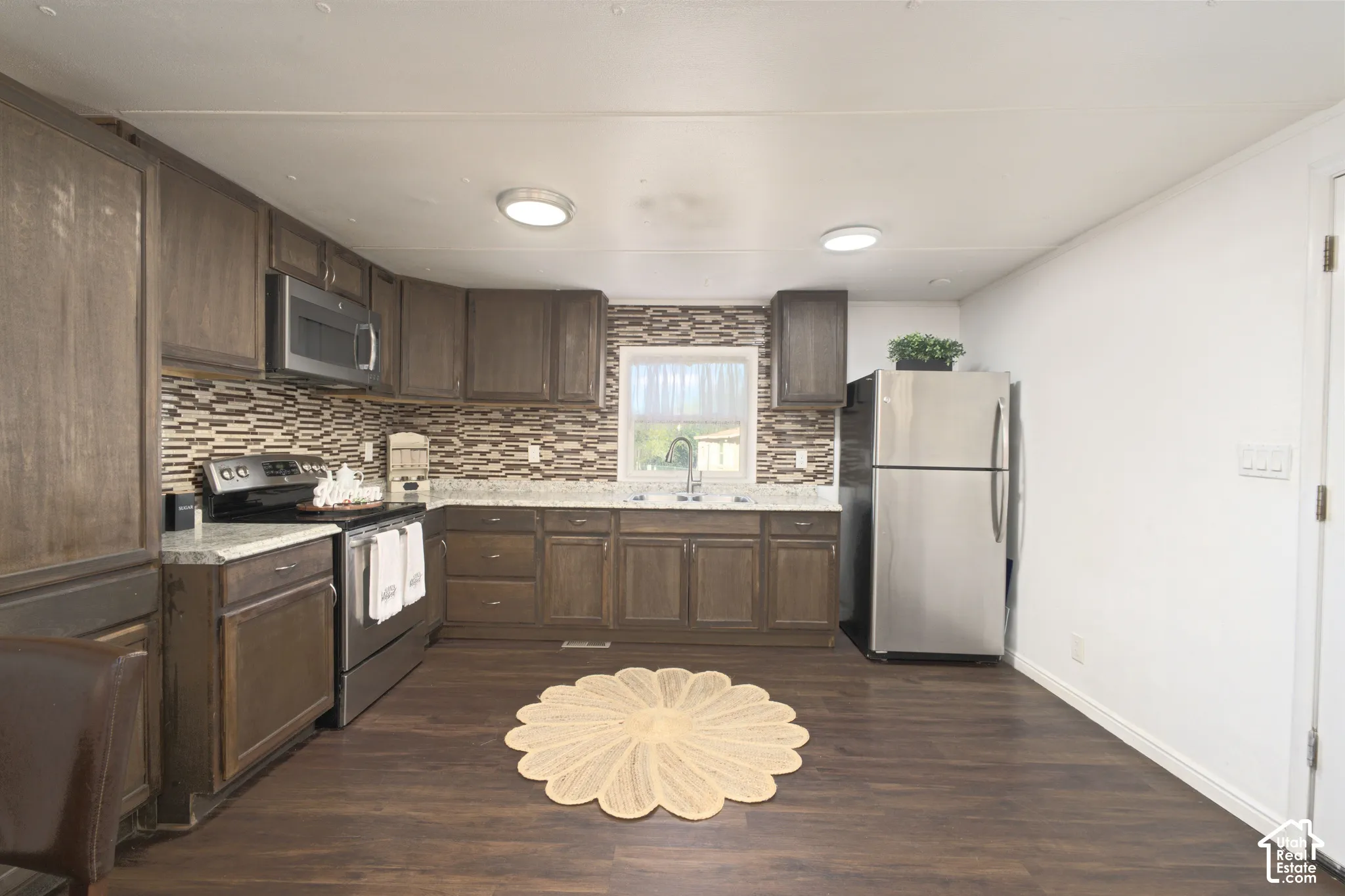 Kitchen featuring dark wood cabinetry, stainless steel appliances, multi-tonal tile backsplash, speckled countertops, and wood-finish flooring