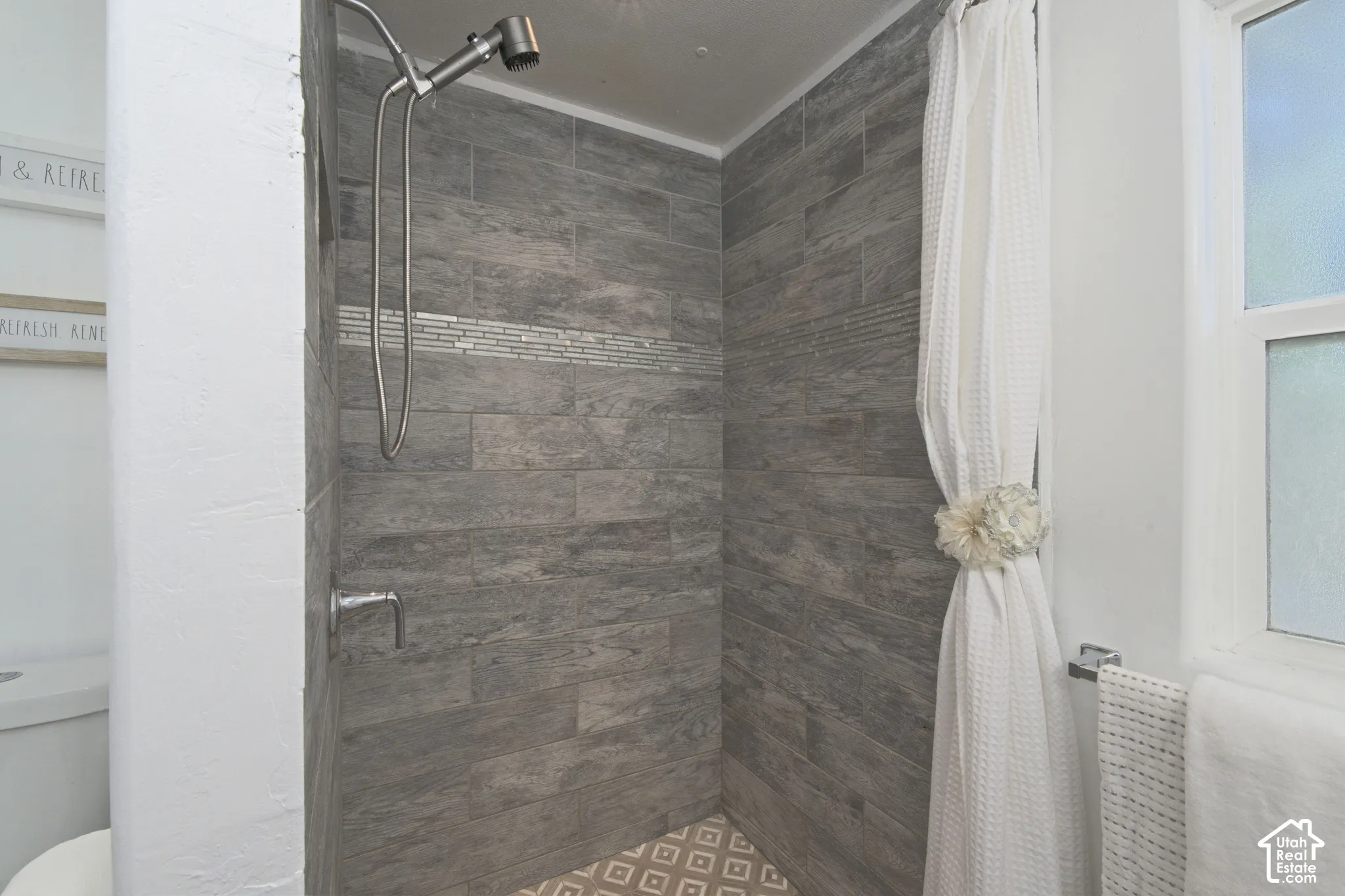 Shower enclosure featuring full-height wood-finish tile surround, integrated decorative accent tile, brushed-finish adjustable shower head, and patterned floor tile