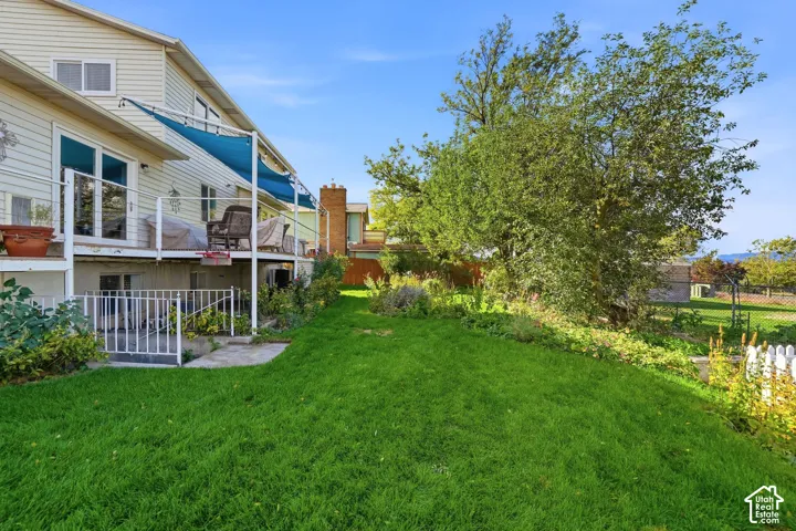 Spacious backyard featuring a manicured lawn, mature trees, elevated metal-railed deck with shade canopy, and metal privacy fencing