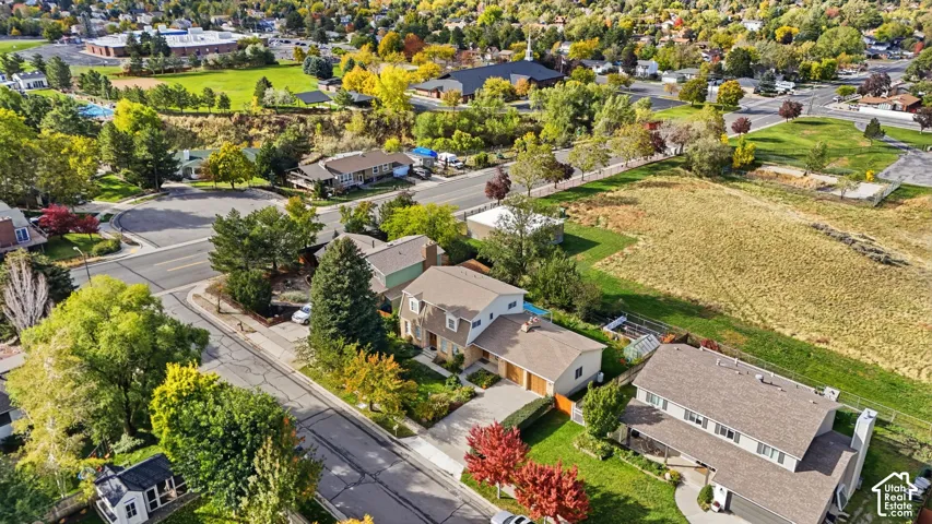 Aerial perspective featuring residential structures with shingled roofs, attached garages, paved driveways, mature trees, and expansive lawn areas