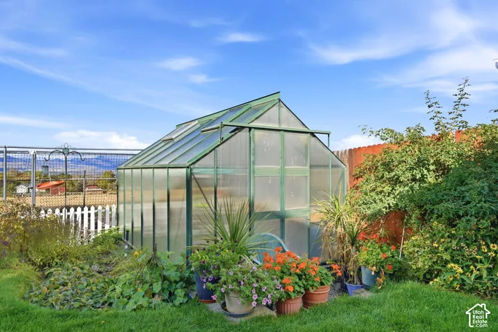Greenhouse featuring a metal-framed structure with translucent panels, adjustable roof vents, and a hinged door, positioned within a landscaped yard