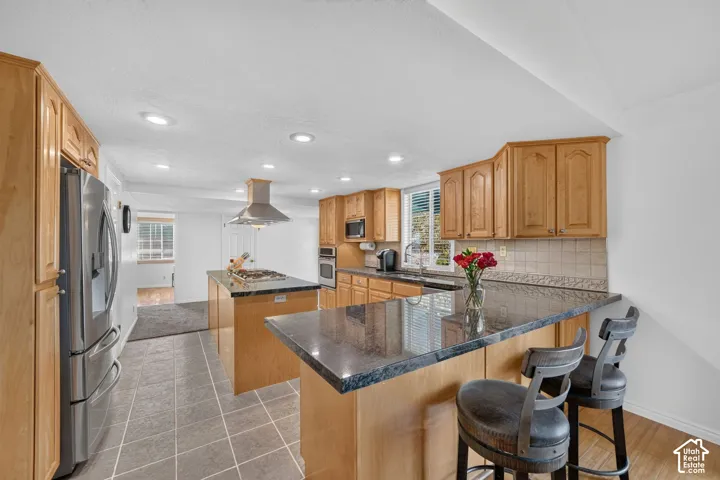 Kitchen featuring light wood cabinetry, stone-finish countertops, stainless steel appliances, a central island with a cooktop and range hood, and recessed lighting