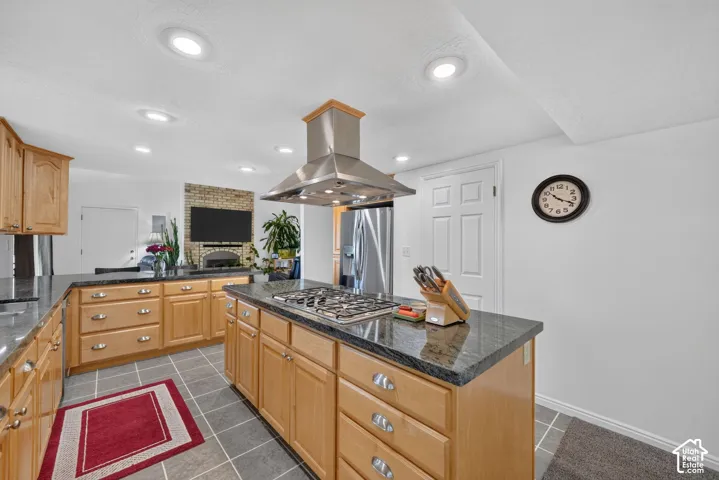 Kitchen featuring light wood-finish cabinetry, dark stone countertops, a central island with a built-in gas cooktop, a stainless steel range hood, and tile flooring