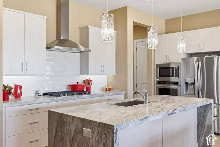 Kitchen featuring light-toned cabinetry, natural stone countertops with waterfall edge detailing, stainless steel appliance suite, white subway tile backsplash, and crystalline pendant lighting