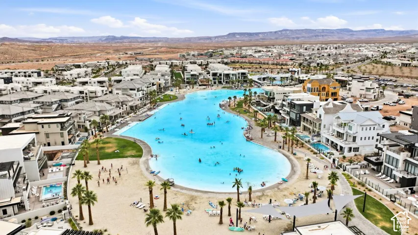 Aerial perspective featuring a large turquoise recreational lagoon, sandy shoreline, mature palm trees, manicured grass areas, and expansive mountain scenery
