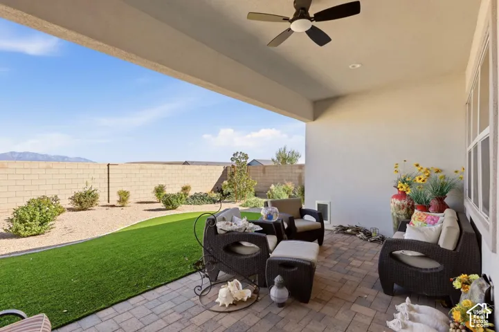Covered patio featuring paver flooring and a ceiling-mounted fan, overlooking a backyard with synthetic turf, desert landscaping, and a masonry perimeter wall