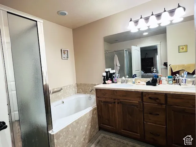 Bathroom featuring a dual-sink vanity with wood cabinetry, a tiled soaking tub deck, a walk-in shower with a frosted glass door, and a full-width wall mirror with integrated multi-bulb vanity lighting