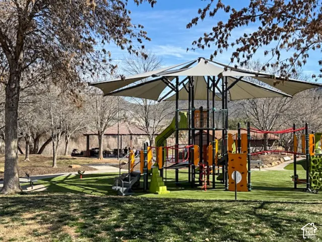 Recreational area featuring a multi-level playground structure with a shade canopy, synthetic turf surfacing, concrete pathways, and a covered pavilion