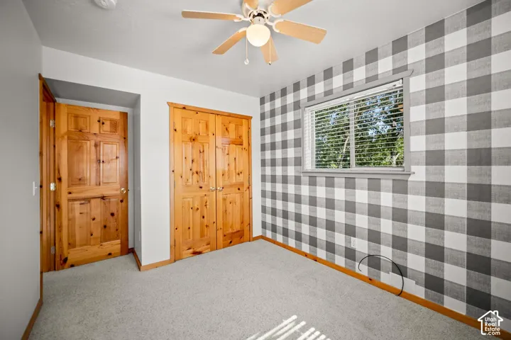 Neutral-toned carpeting, accent wall with gingham pattern, natural wood-finish closet doors, interior door with natural wood finish, and a ceiling-mounted fan with light fixture