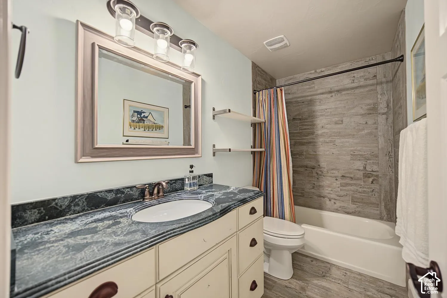 Bathroom vanity with a stone countertop, oval undermount sink, three-light fixture, floating wall shelving, and tub-shower combination featuring horizontal stone-finish wall tile