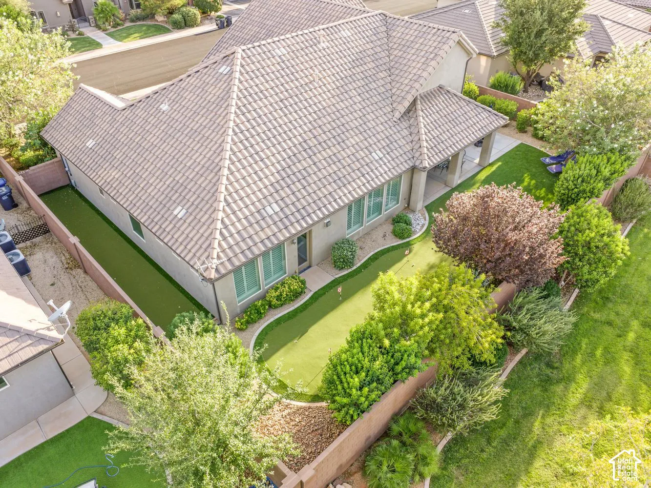 Backyard featuring a synthetic turf putting green, covered patio with structural columns, concrete hardscaping, perimeter masonry wall, and mature landscape plantings