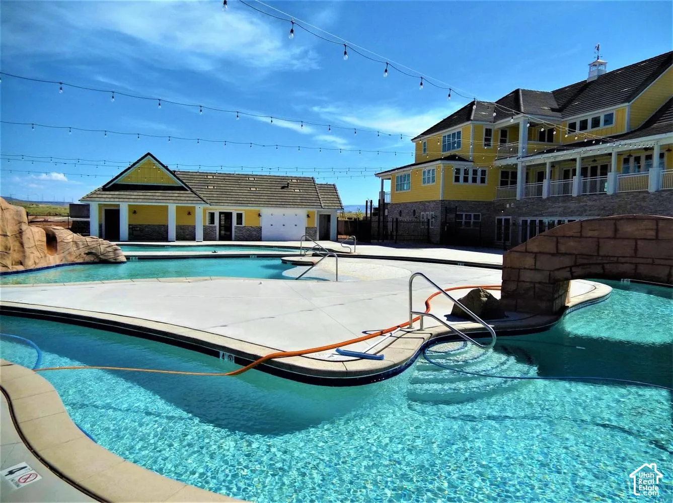 Expansive swimming pool with an integrated stone bridge, rock formation, concrete decking, and overhead string lighting