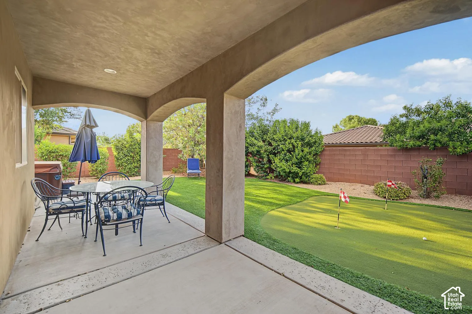 Covered concrete patio featuring arched architectural details, synthetic turf putting green, manicured lawn, and masonry perimeter wall