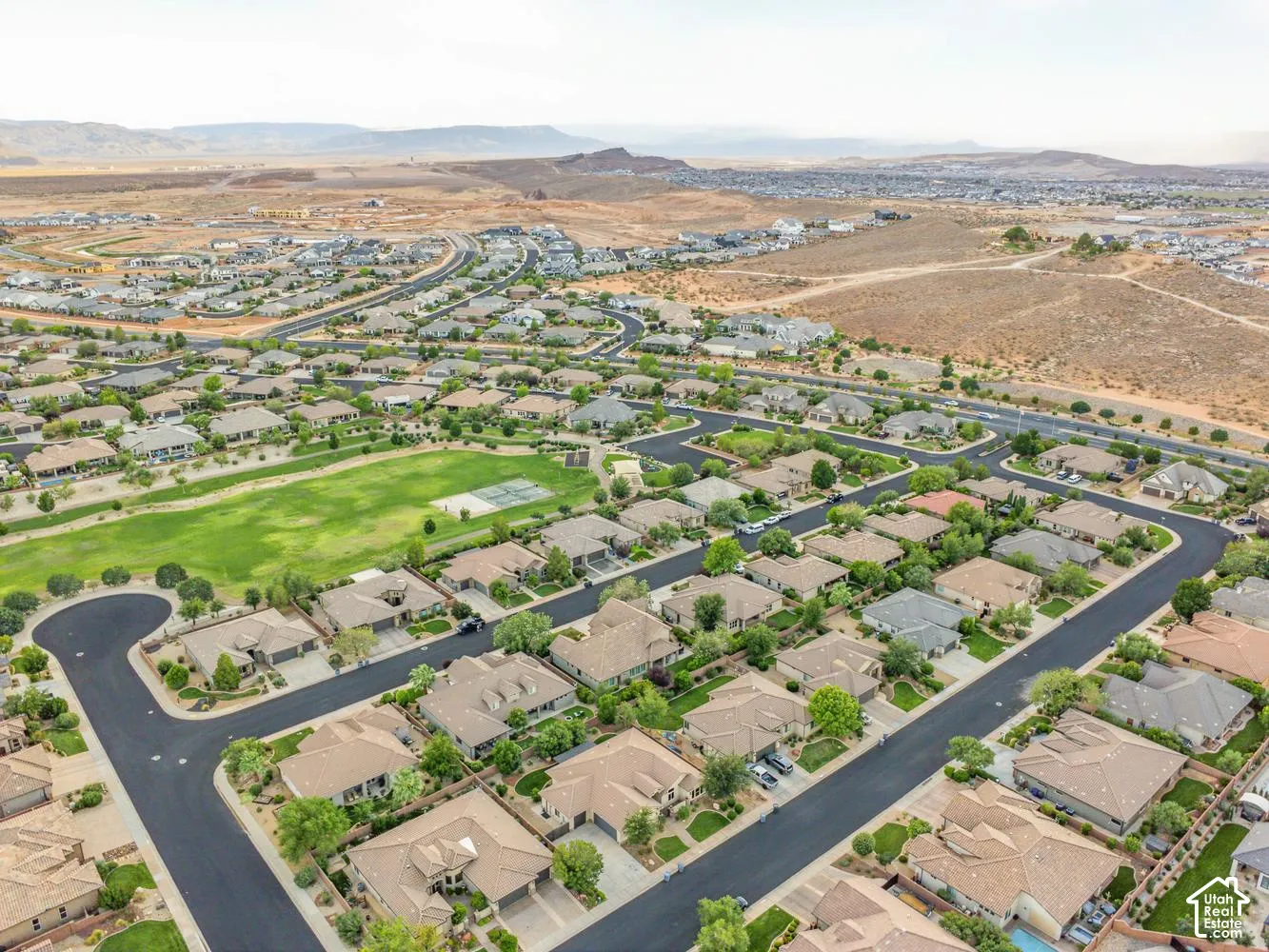 Aerial perspective of a planned community featuring paved asphalt streets, single-story residential architecture, expansive green space, and a mountainous desert horizon