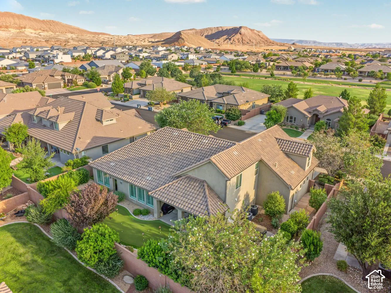 Aerial perspective of a multi-story residence featuring a tile roof, covered rear patio, integrated putting green, and enclosed yard with mature landscaping, set against a backdrop of desert mesas