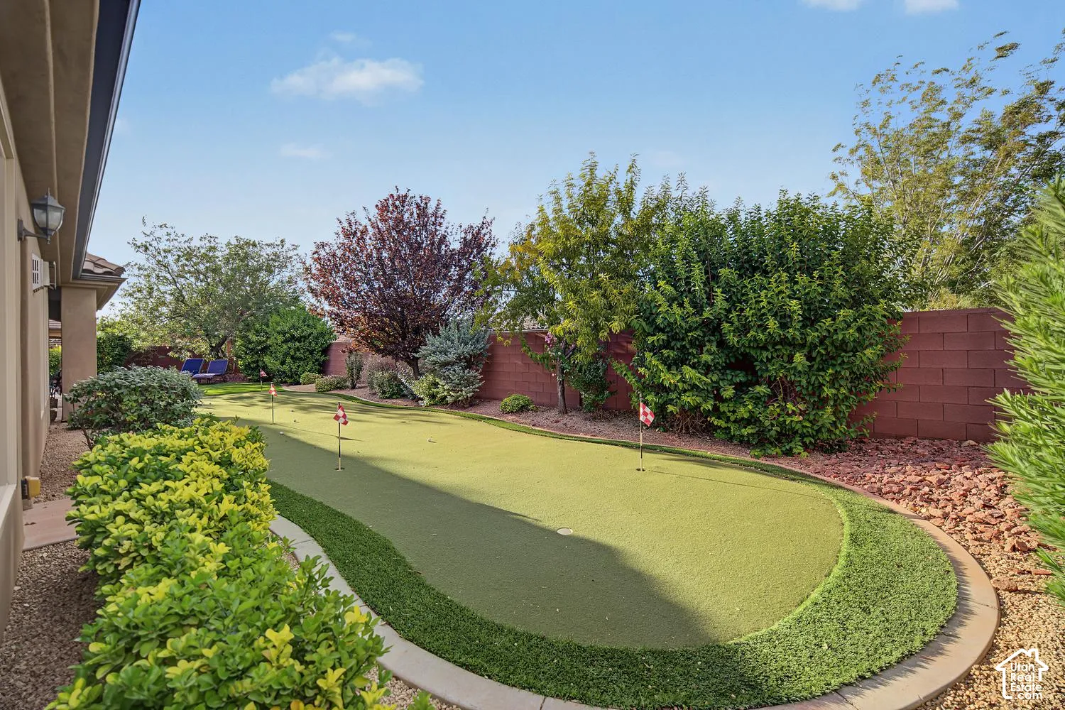 Backyard putting green featuring synthetic turf, multiple pins, and professional concrete edging, bordered by mature landscaping and a masonry privacy wall