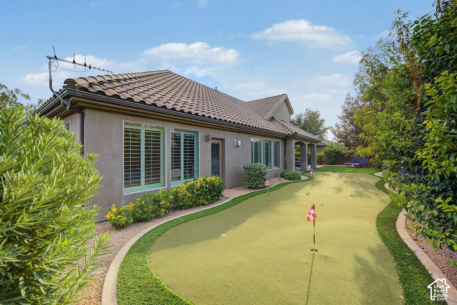 Backyard putting green featuring artificial turf, multiple pins, and concrete curbing, accented by mature landscaping and a stucco exterior home with a tiled roof