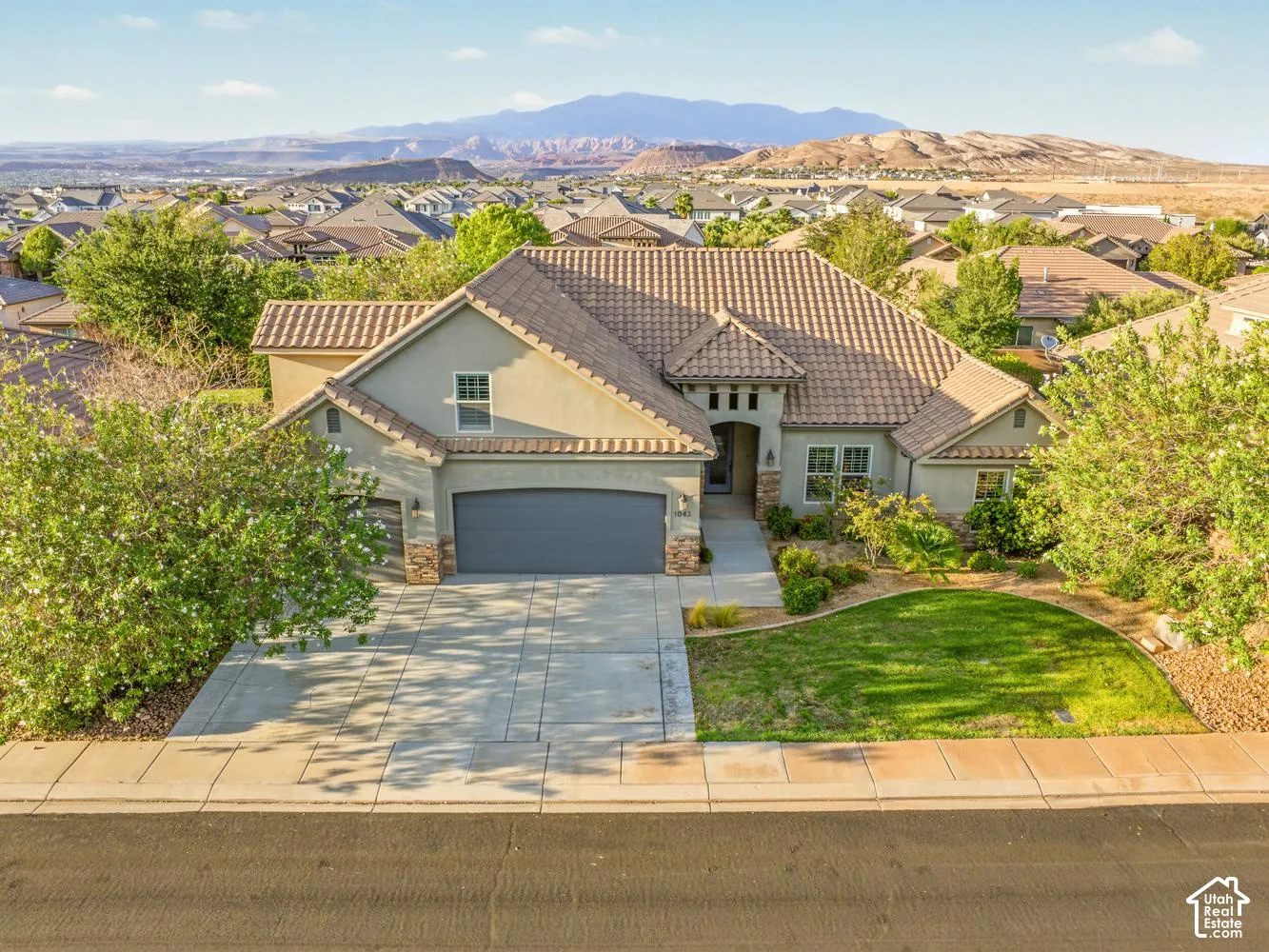 Stucco exterior featuring a tile roof, multi-car garage, concrete driveway, and established landscaping with mountain backdrop