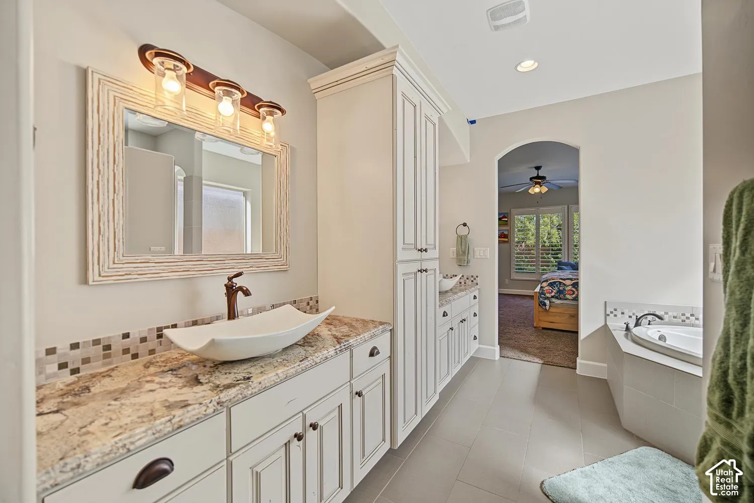 Bathroom featuring a custom vanity with stone-surfaced countertops, a vessel sink, tall built-in cabinetry, and a jetted soaking tub with tile surround