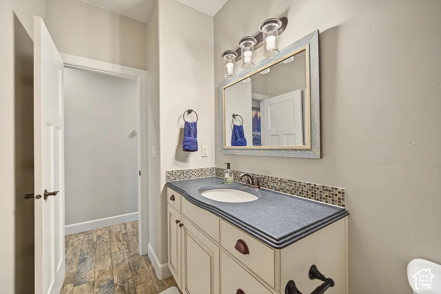 Bathroom featuring a vanity with a custom-edged countertop, undermount sink, multi-tone mosaic tile backsplash, wood-finish flooring, and a framed mirror with three-light fixture